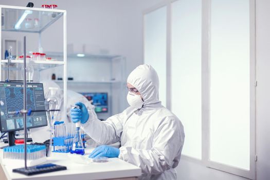 Man scientist filling micropipette with solution from glass flask dressed in coverall. Chemist in modern laboratory doing research using dispenser during global epidemic with covid-19.