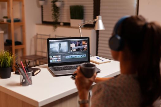 Videographer holding cup of coffee while working in video footage during post production. Content creator in home working on montage of film using modern software for editing late at night.