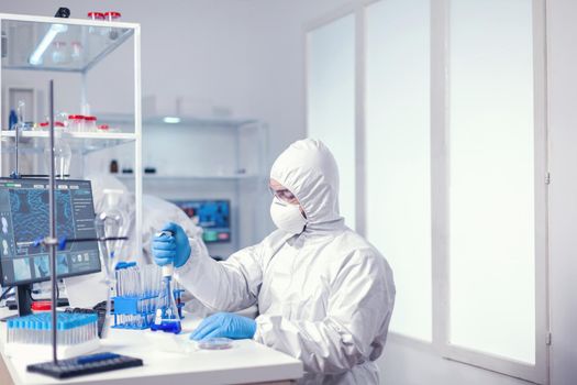 Medical worker uses micropipette in modern scientific facility wearing protection suit as safety precaution. Chemist in modern laboratory doing research using dispenser during global epidemic with covid-19.