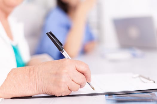 Mature woman doctor taking notes on clipboard during briefing with coworkers. Clinic expert therapist talking with colleagues about disease, medicine professional