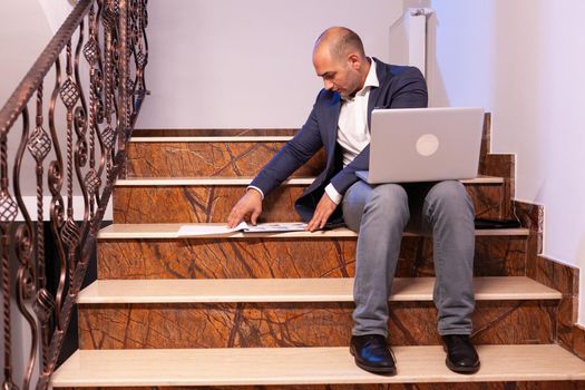 Stressed tired businessman at night in workplace finishing project deadline. Confident corporate entrepreneur using laptop doing overtime sitting on stairwell.