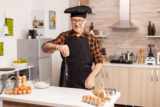 Elderly man preparing food spreading flour in home kitchen for food preparation. Retired senior chef with bonete and apron, in kitchen uniform sprinkling sieving sifting ingredients by hand.