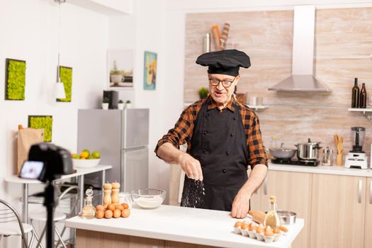 Experienced senior chef recording tutorial with food preparation in kitchen. Retired blogger baker influencer using internet technology communicating, shooting, blogging on social media with digital equipment