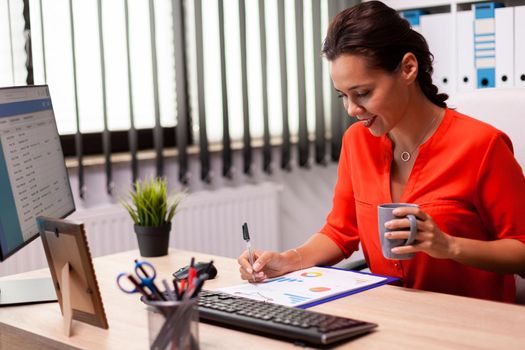 Professional businesswoman entrepreneur reading finance analysis looking at corporate charts wearing red. Successful financial auditor in data marteking looking at graphs on computer screen and writing report.