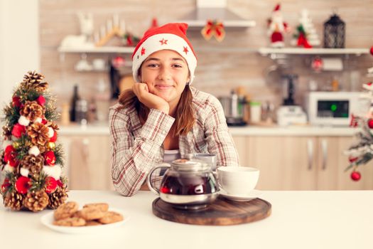 Pensive child enjoying chritmas and tasty cookies on kitchen table and christmastree. Cheerful happy adorable teenager girl in home kitchen with delicious biscuits and xmas tree in the background celebrating winter holiday.