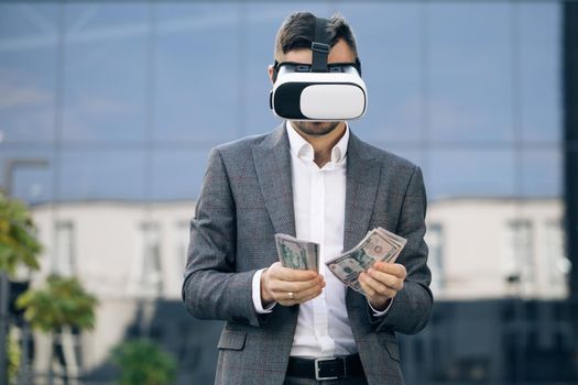 Rich man wearing virtual reality headset counting money. Young businessman standing with pack of dollars near modern office building. Richness and success concept. 3D VR Glasses.