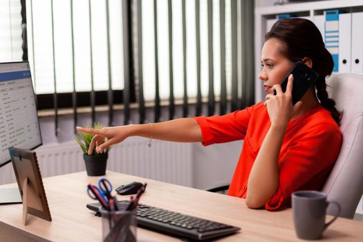 Entrepreneur businesswoman in professional call pointing at documents on computer screen. Busy freelancer working using smartphone from office to talk with clients sitting at desk looking at document.