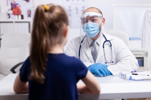 Doctor wearing visor for safty precaution during coronavirus while having child for examination. Specialist in medicine providing health care services consultation, radiographic treatment in clinic cabinet hospital