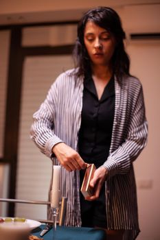 Wife lighting candle in kitchen for romantic dinner with husband. Caucasian happy woman preparing festive meal with healthy food for anniversary celebration, table in kitchen.