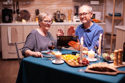 Smiling senior couple waving at camera of tablet pc during video call and dinner in kitchen. Couple sitting at the table, browsing, talking, using internet, celebrating their anniversary in dining room.