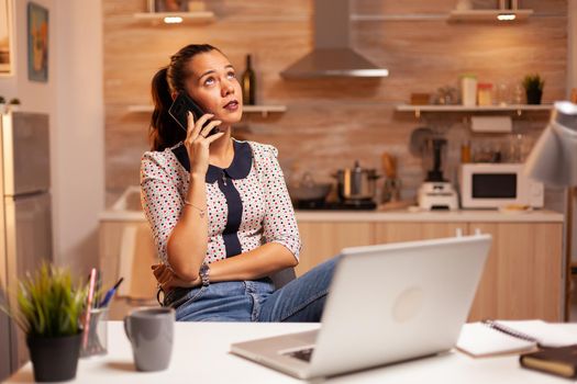 Freelance woman having a phone coversation with a client late at night from home kitchen. Employee using modern technology at midnight doing overtime for job, business, career, network, lifestyle.