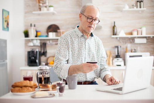 Senior man doing online purchase in morning from his kitchen. Pensioner paying online using credit card and application from laptop. Retired elderly person using internet payment home bank buying with modern technology