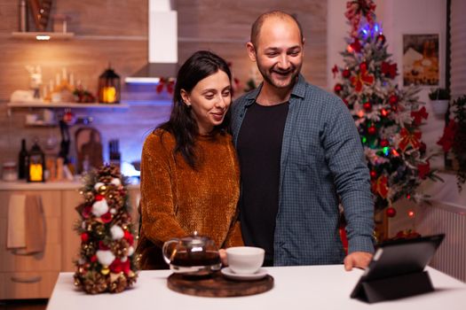 Joyful couple discussing with remote friends during online videocall meeting conference using laptop computer standing at table in xmas decorated kitchen. Haapy family celebrating christmas holiday