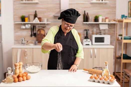 Senior lady chef smiling while preparing pizza sprinkling flour on kitchen table. Happy elderly chef with uniform sprinkling, sieving sifting raw ingredients by hand.