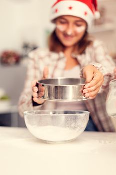 Girl sifts flour for tasty cookies for christmas. Happy cheerful joyfull teenage girl helping senior woman preparing sweet biscuits to celebrate winter holidays wearing santa hat.