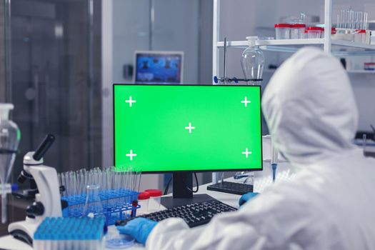 Over shoulder shot of doctor working on coroanvirus vaccine using computer with green screen dressed in ppe. Team of microbiologists doing vaccine research writing on device with chroma key, isolated, mockup display.