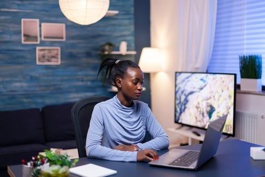 Dark skinned freelancer reading documentation on laptop computer in living room late at night. Black entrepreneur sitting in personal workplace writing on keyboard.