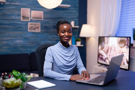 Dark skinned business woman using digital device working late at night in home office. Black entrepreneur sitting in personal workplace writing on keyboard.