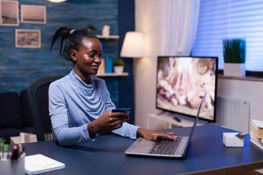 Dark skinned business woman holding credit card doing financial purchase for business from home office. Employee making paymant transaction from home on digital notebook.