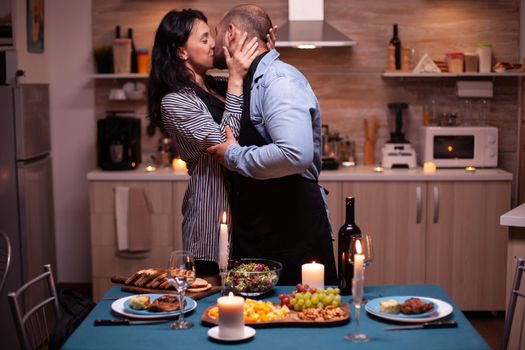 Husband kissing delighted wife while celebrating their relationship in kitchen with tasty food. Man preparing festive dinner with healthy food, cooking for his woman a romantic dinner,