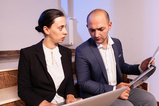 Businesswoman doing overtime during late night hours with coworker for difficult deadline. Colleague entrepreneur working late together in evening at corporate job sitting on staircase, explaining difficult finance project.