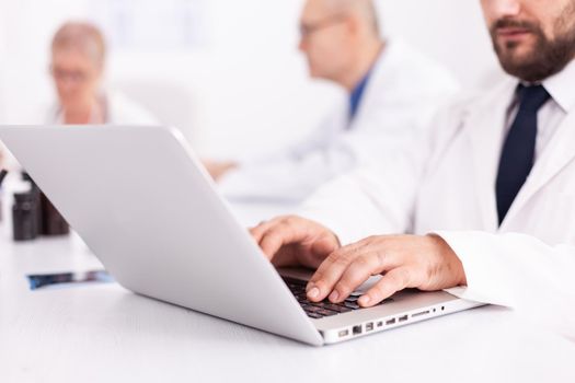 Young medical specialist typing on laptop during meeting with coworkers in hospital office. Clinic expert therapist talking with colleagues about disease, medicine professional