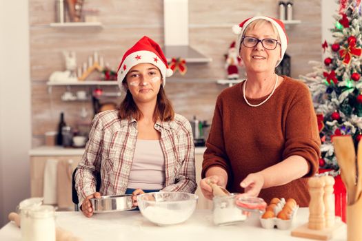 Niece and grandmother smiling looking at camera during christmas celebration .Happy cheerful joyfull teenage girl helping senior woman preparing sweet cookies to celebrate winter holidays wearing santa hat.