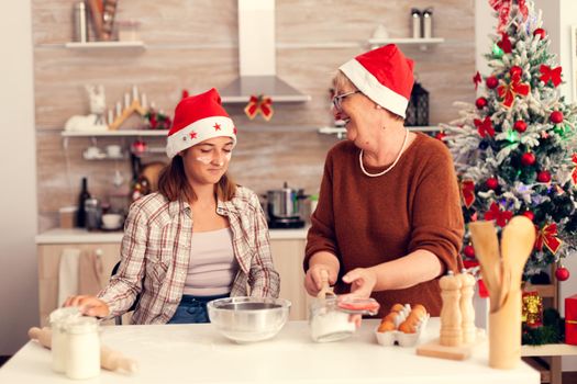 Grandmother and granddaughter on christmas day baking biscuits. Happy cheerful joyfull teenage girl helping senior woman preparing sweet cookies to celebrate winter holidays wearing santa hat.