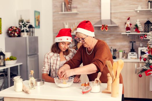 Grandmother cracking eggs over flour on christmas day. Happy cheerful joyfull teenage girl helping senior woman preparing sweet cookies to celebrate winter holidays wearing santa hat.