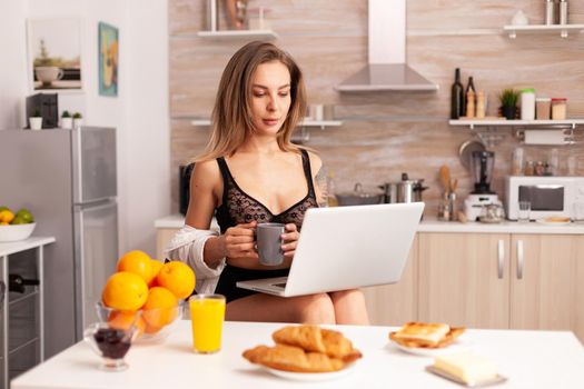 Attractive blonde woman in sexy lingerie holding coffee while using laptop in home kitchen. Beautiful lady with tattos during breakfast in underwear.