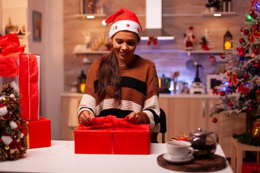 Caucasian woman preparing gift boxes with ribbon and wrapping paper for christmas eve celebration party. Festive young person wirh santa hat in seasonal holiday decorative kitchen