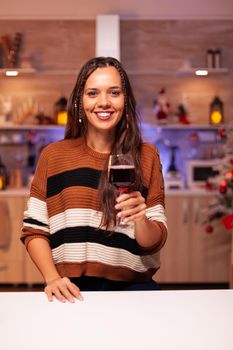 Portrait of cheerful woman holding glass of wine in festive kitchen decorated for christmas eve dinner celebration. Caucasian young person enjoying drink in winter season at home