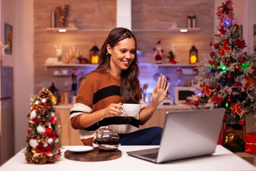 Young woman holding cup of tea using video call concept for online conference on laptop in festive kitchen at home. Caucasian adult enjoying chat with relatives on christmas eve night