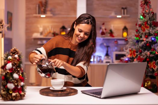 Festive person using video call technology on laptop while pouring cup of tea enjoying winter season at home. Caucasian young woman talking to family online in christmas decorated room