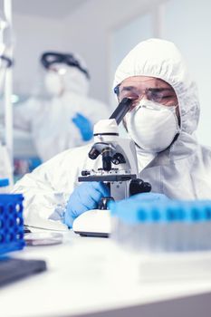 Medical researcher wearing coverall searching for covid treatment looking through microscope. Scientist in protective suit sitting at workplace using modern medical technology during global epidemic.