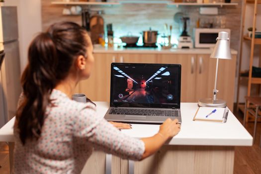 Female game developer working on new project on personal laptop in home kitchen during night time. Professional gamer playing online videop games on her personal computer. Geek cyber e-sport.
