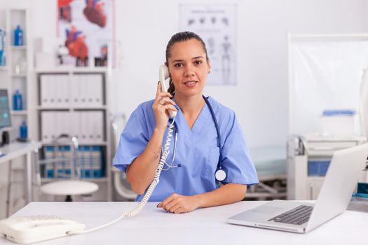 Portrait of medical nurse smiling at camera while using phone in hospital office. Health care physician sitting at desk using computer in modern clinic looking at monitor.