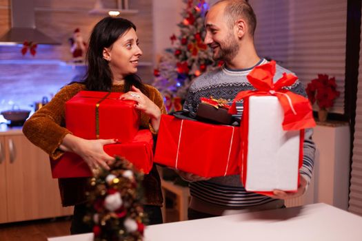 Happy married couple holding secret present gift with ribbon on it during christmas holiday standing in xmas decorated kitchen. Smiling family enjoying winter season celebrating christmastime together