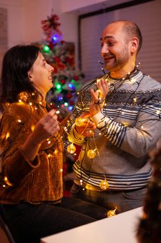 Amused couple having fun with christmas tree light decorating kitchen enjoying spending wintertime together. Happy family celebrating santa-claus festive season. Christmastime tradition