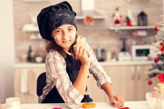 Cheerful grandchild on christmas day holding heart shaped pastery wearing apron and bonette. Happy joyfull teenage girl helping senior woman preparing sweet cookies to celebrate winter holidays.