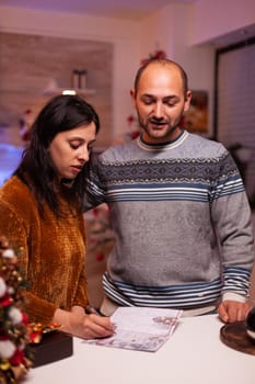 Cheerful girlfriend writing christmas greeting postcard celebrating xmas winter season with boyfriend standing in x-mas decorated kitchen, Happy family enjoying spending christmastime together