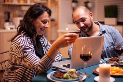 Woman holding credit card while doing shopping with husband during dinner. Adults sitting at the table, searching, browsing, surfing, using technology card payment, internet