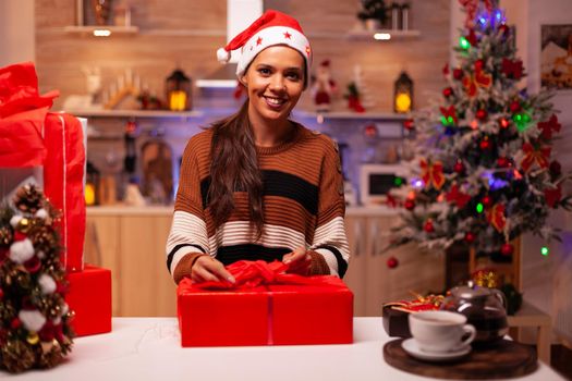 Caucasian woman preparing gift boxes with ribbon and wrapping paper for christmas eve celebration party. Festive young person with santa hat in seasonal holiday decorative kitchen