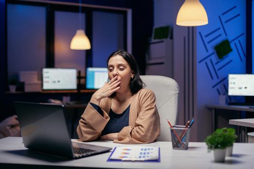 Sleepy businesswoman yawns while working on laptop trying to finish a project deadline. Smart woman sitting at her workplace in the course of late night hours doing her job.