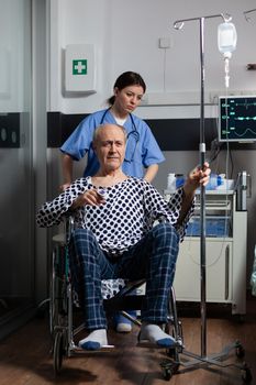 Hospitalized senior man sitting in wheelchair in hospital room, holding iv drip with oximeter attached on finger. Medcal assistant preparing sick patient for treatment.