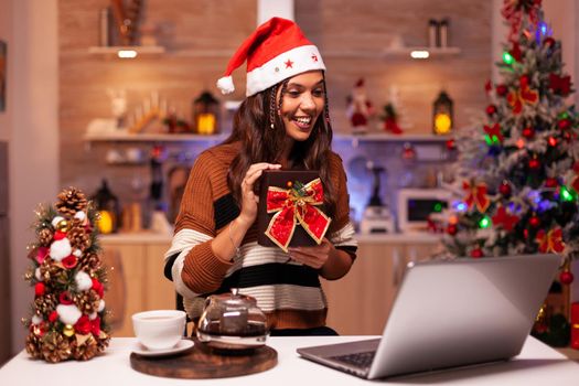 Caucasian woman opening present on video call conference technology with friends in festive home. Cheerful young person receiving surprise gift for christmas eve seasonal celebration