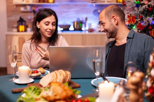 Happy family shopping xmas present gift using credit card doing online payment on laptop computer sitting at x-mas table in kitchen. Cheerful couple celebrating christmas holiday