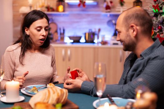 Haapy man proposing girlfriend to marry him using diamond luxury expensive engagement ring celebrating christmas holiday enjoying winter season together. Cheerful couple sitting at xmas dinning table