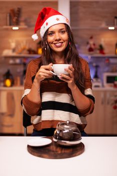 Portrait of young woman wearing santa hat and smiling in kitchen decorated with ornaments, lights and tree for christmas eve. Caucasian adult thinking about winter celebration festivity