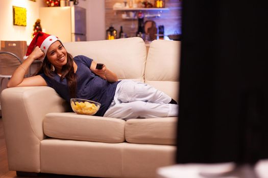 Caucasian female resting on sofa changing channel using remote control looking at christmas movie on television. Woman wearing red santa hat celebrating winter holiday enjoying christmastime